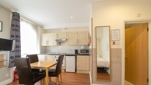 A compact kitchen with wooden cabinets, a microwave, and a small refrigerator. There is a dining table with chairs and a fire extinguisher near the entrance. - Imperial Court Studios (London)