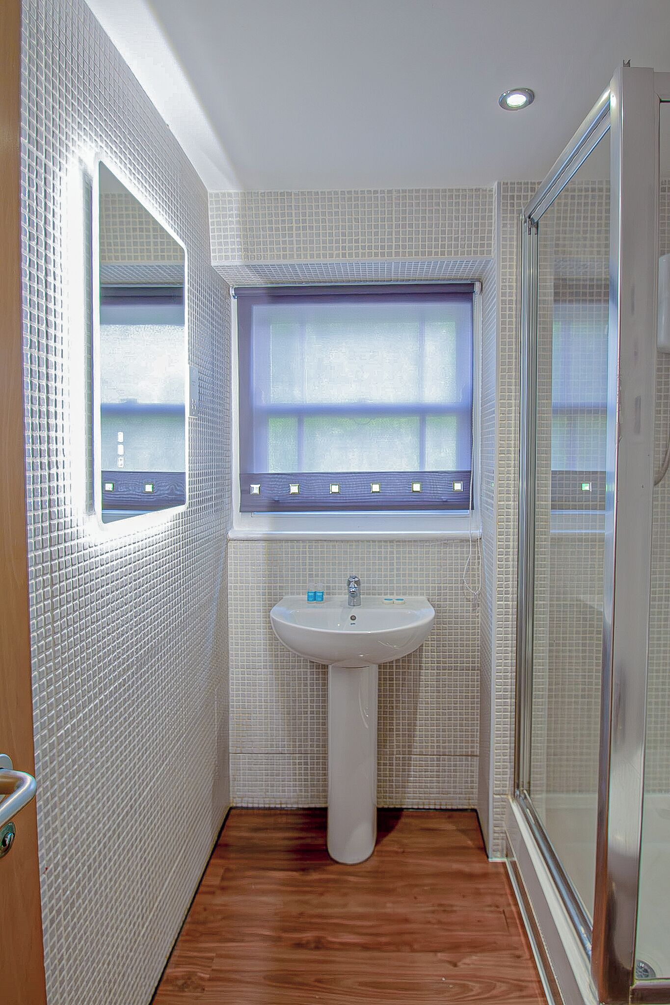 A bathroom with a white tiled wall, a window with blinds, a white sink, and a wooden floor.