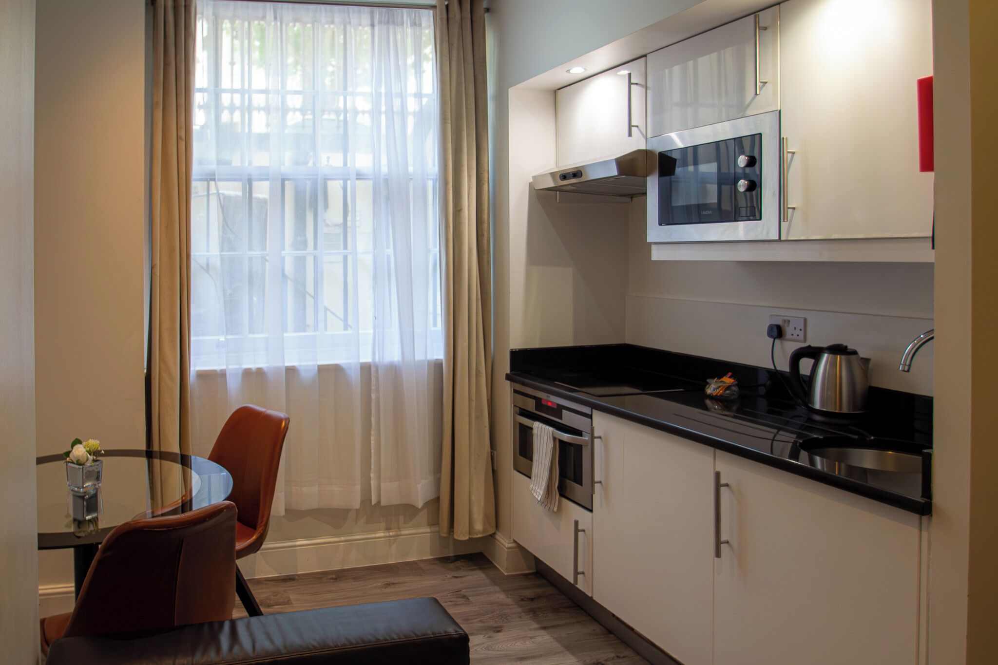 A compact kitchen with a microwave, oven, and kettle, a dining table with chairs, and a window with sheer curtains.