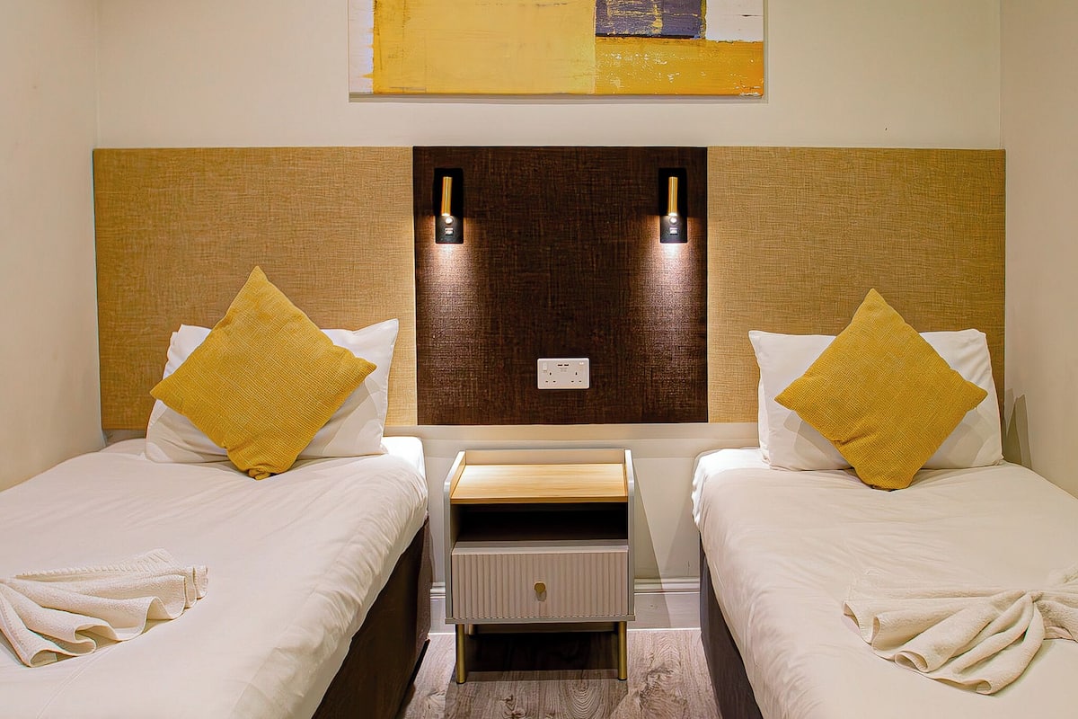 Een hotelkamer met twee bedden, elk met een witte matras en een geel kussen. Tussen de bedden staat een houten nachtkastje met een lade.