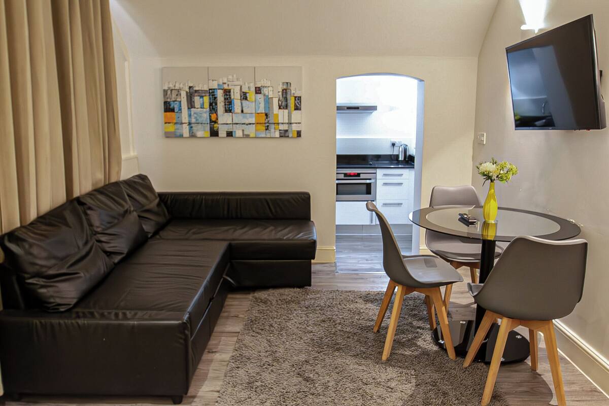 Een moderne woonkamer met een zwarte leren bank, een ronde glazen tafel met stoelen, een kleine vaas met bloemen en een flatscreen televisie aan de muur.