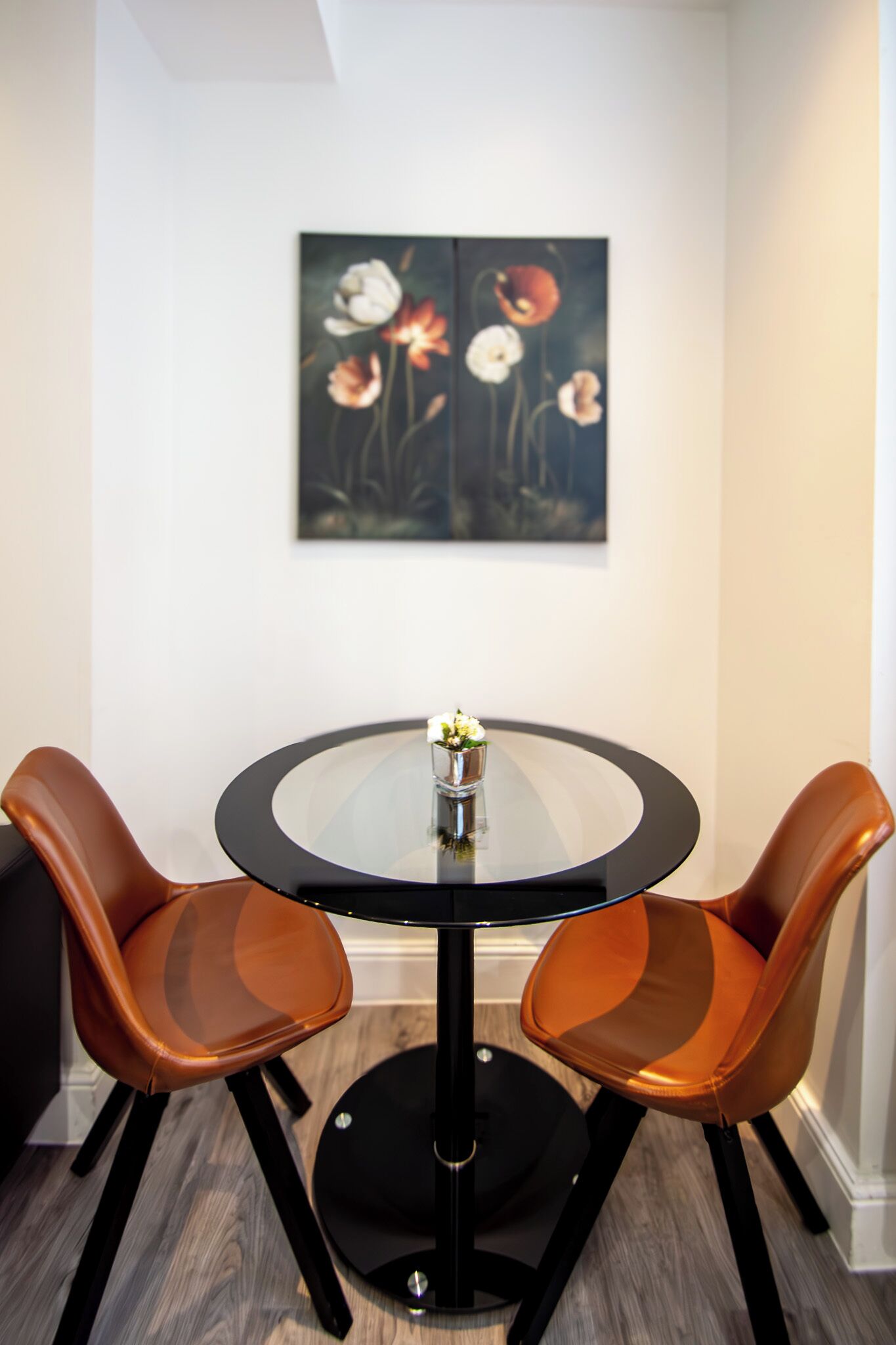 Une table ronde en verre, accompagnée de deux chaises en cuir marron, un petit vase avec des fleurs blanches et un tableau représentant des fleurs accroché au mur.