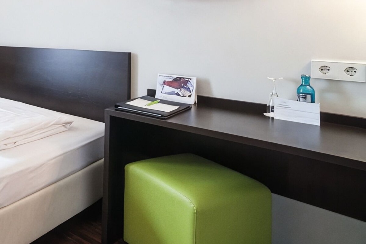 Ein Hotelzimmer mit einem Bett, einem Schreibtisch, einem Stuhl und einem grünen Hocker.