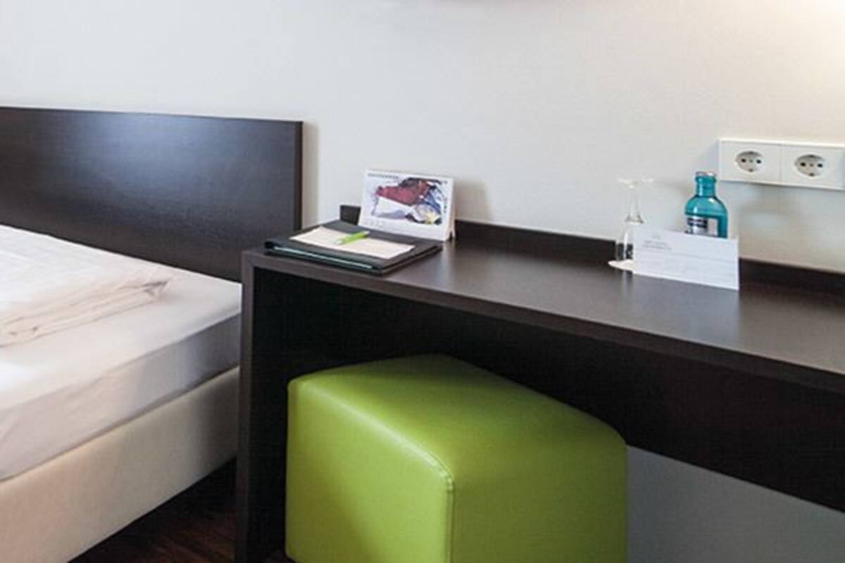 Quarto de hotel com uma cama, uma escrivaninha, uma cadeira e um banquinho.