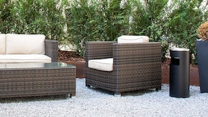 An outdoor seating area with wicker furniture and a glass table, surrounded by gravel and greenery.