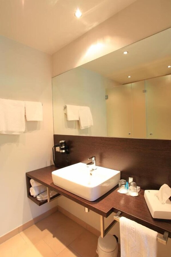 Un baño moderno con un espejo grande, un lavabo blanco y un mueble de tocador de madera. Hay toallas colgadas en la pared y un cubo de basura.
