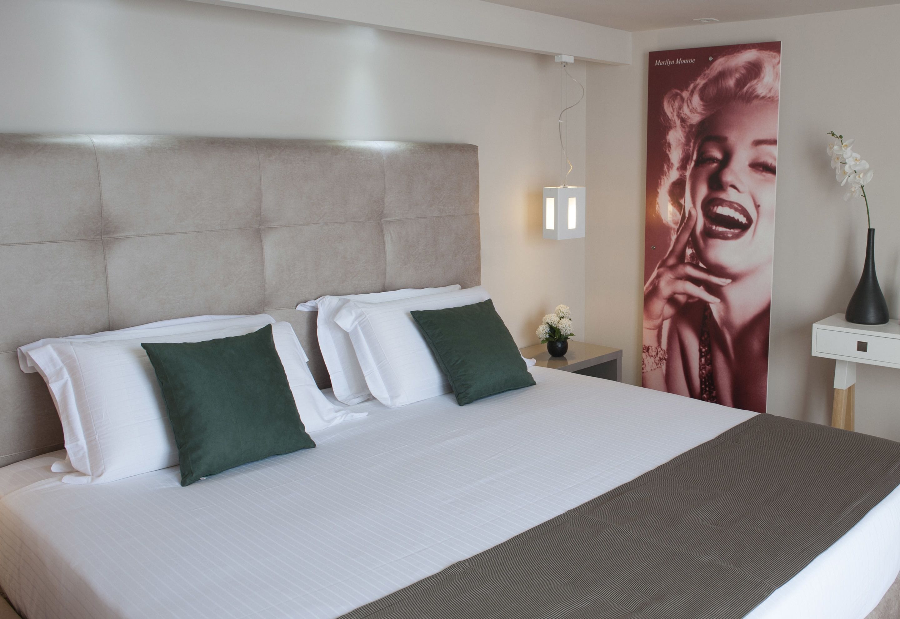 Ein ordentlich bezogenes Bett mit weißen und grünen Kissen, einem grauen Kopfteil und einem Marilyn-Monroe-Poster an der Wand.
