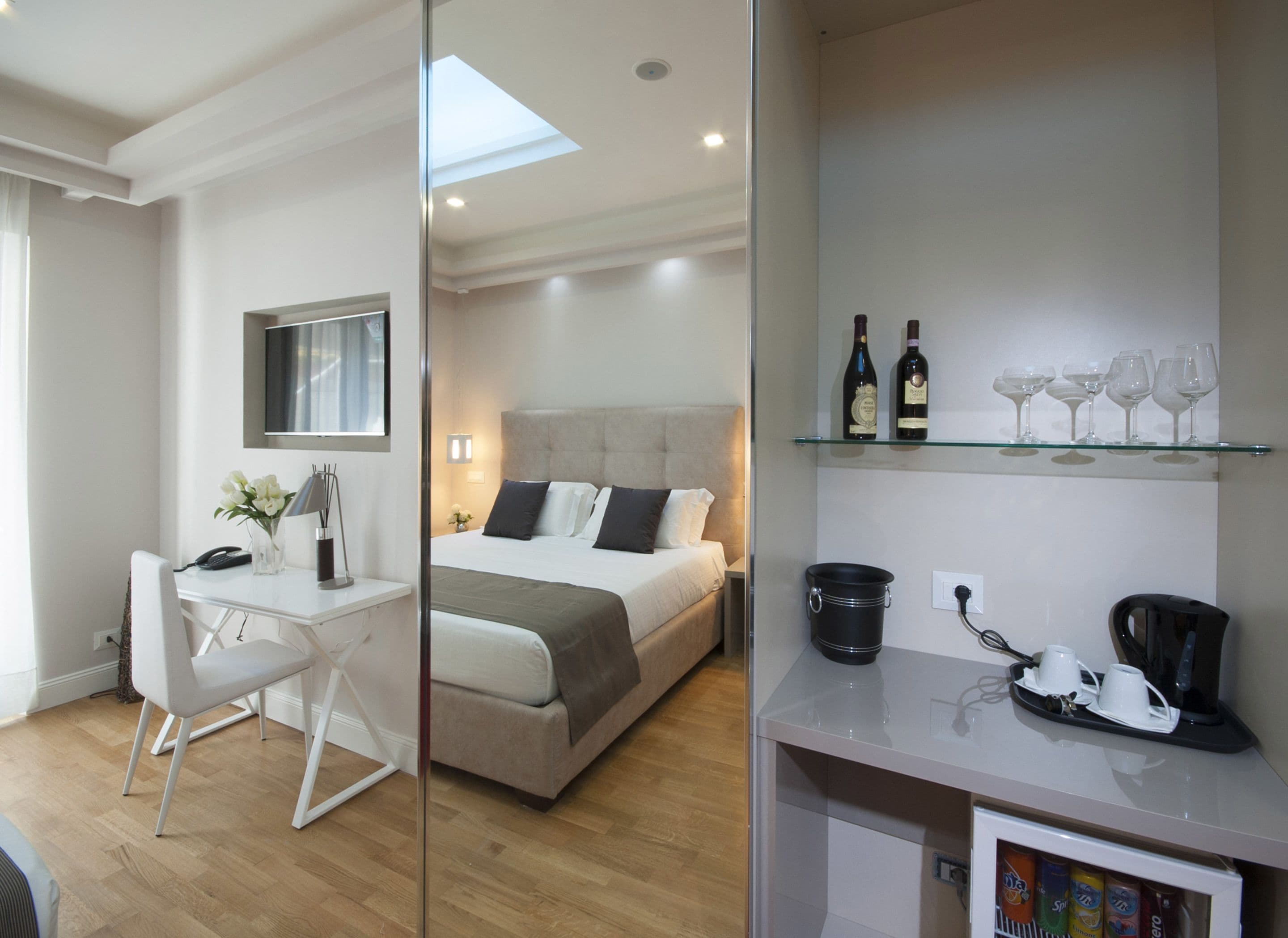 A modern hotel room with a bed, a desk, a chair, a small fridge, and a shelf with wine bottles and glasses.