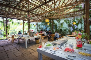 Un área de comedor al aire libre con mesas y bancos de madera, rodeada de exuberante follaje y una estructura de techo de bambú.