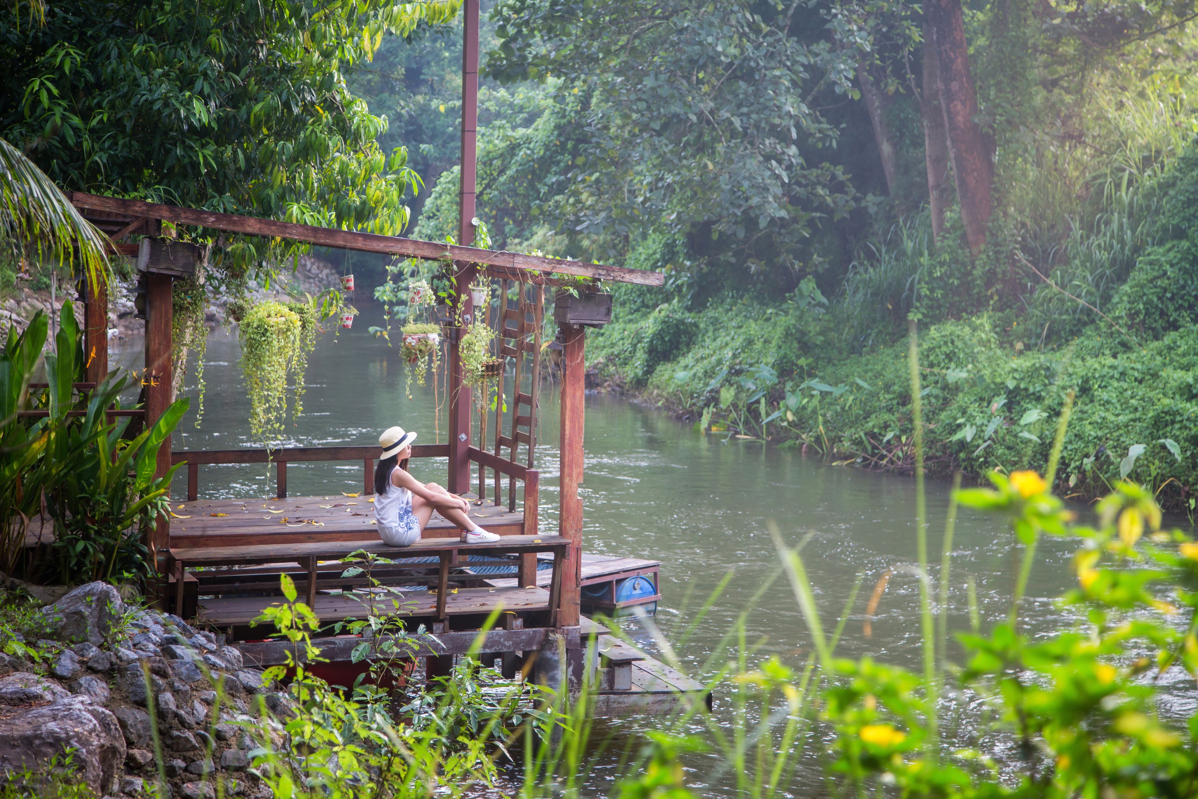 A wooden dock with a person sitting, surrounded by lush greenery and a calm river.