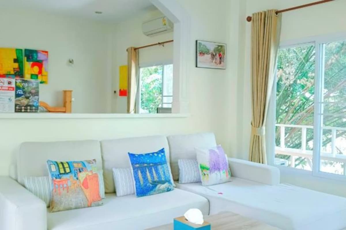 A white sofa with colorful cushions, a small table with a lamp, and a window with curtains.