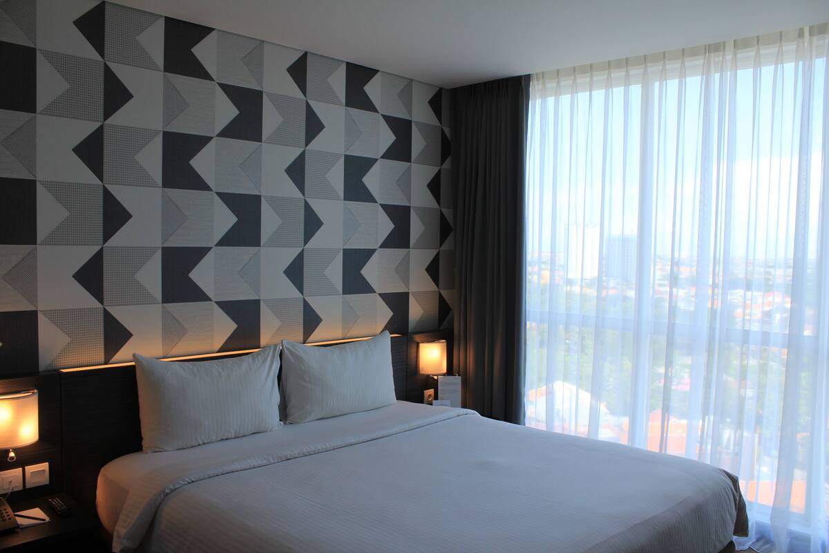 A hotel room with a large bed, two bedside tables with lamps, a geometric patterned wall, and a view through sheer curtains.