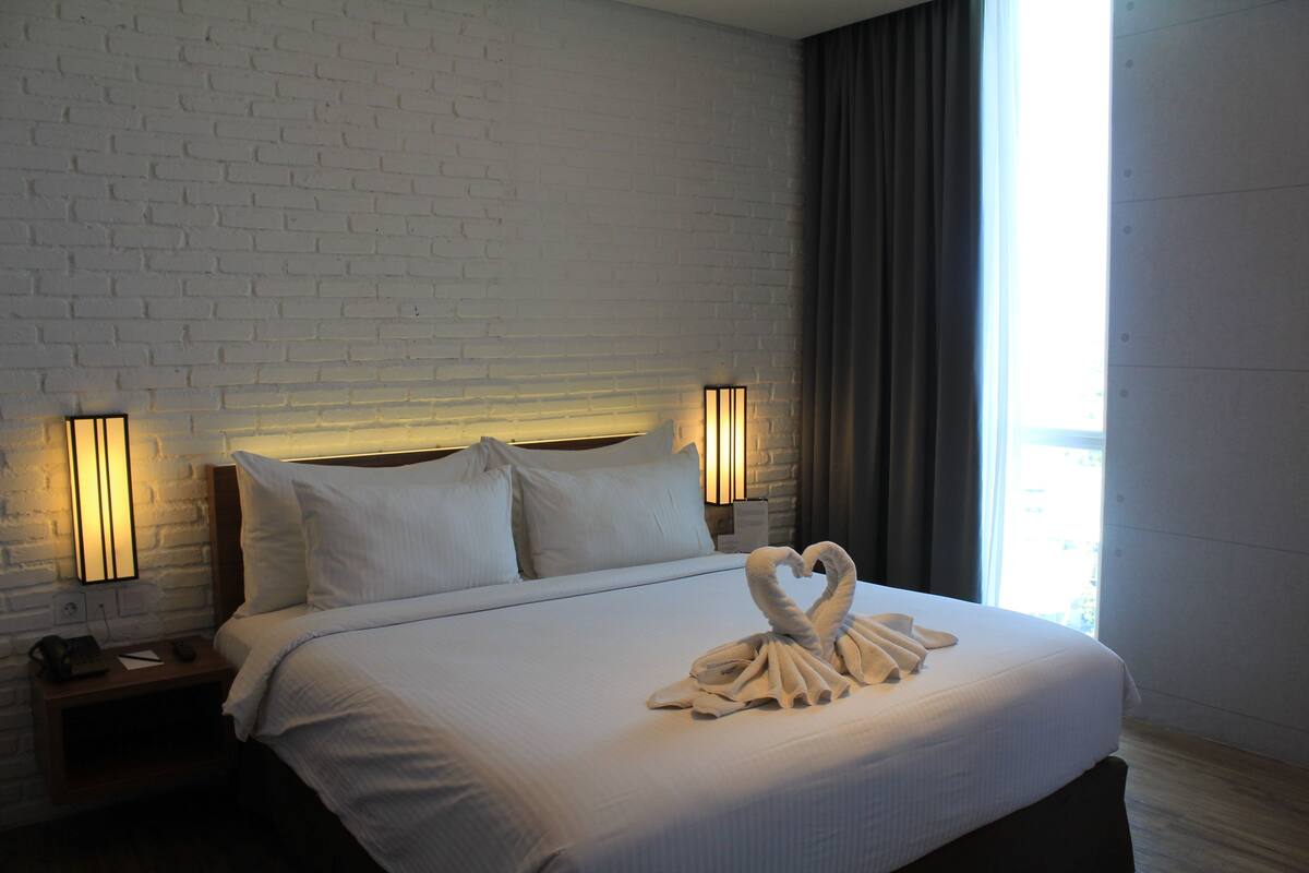 A hotel room with a white brick wall, a large bed with white linens, two bedside lamps, and a heart-shaped towel arrangement on the bed.
