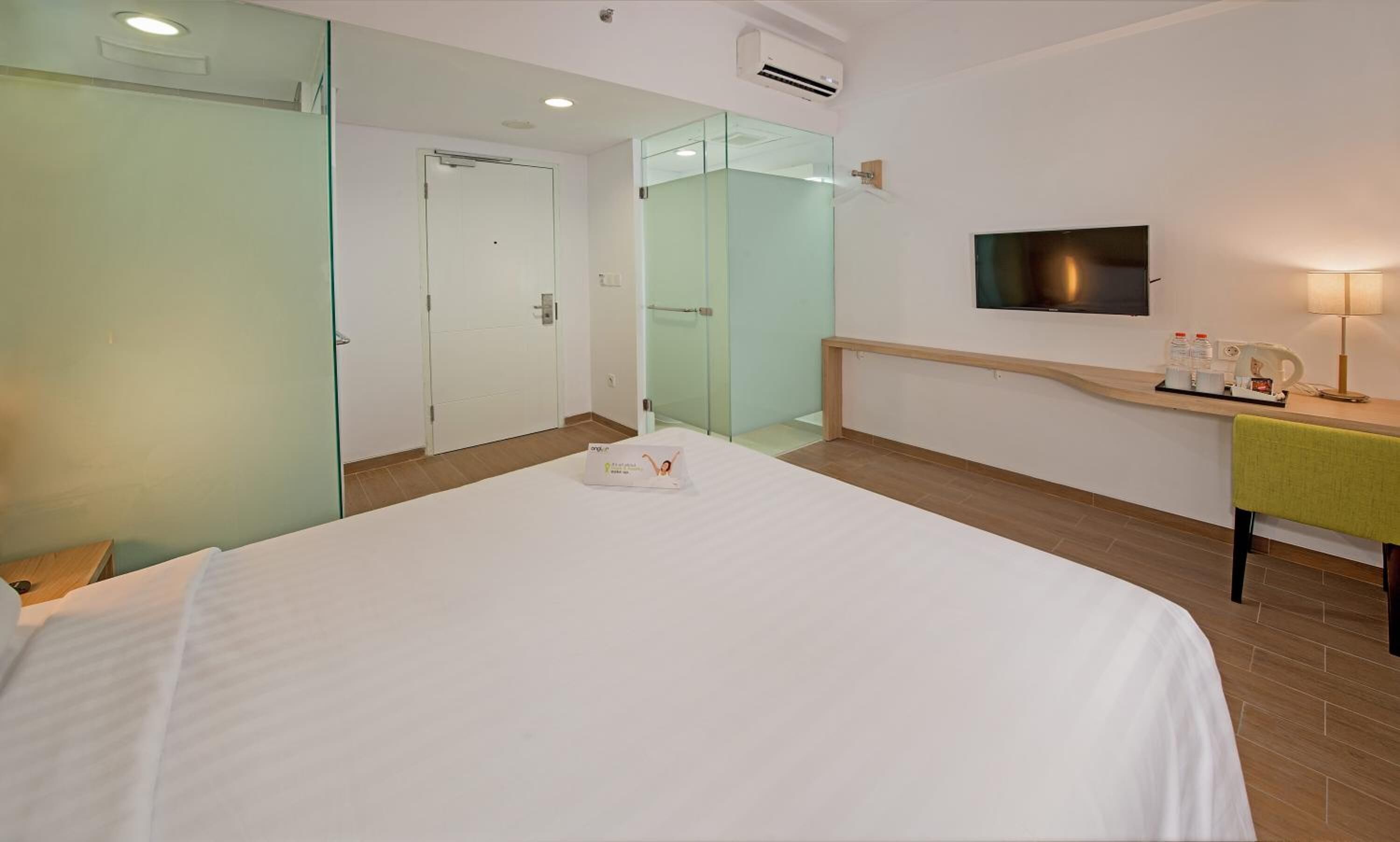 A hotel room with a bed, a desk with a lamp, a TV, and a glass door leading to a bathroom.