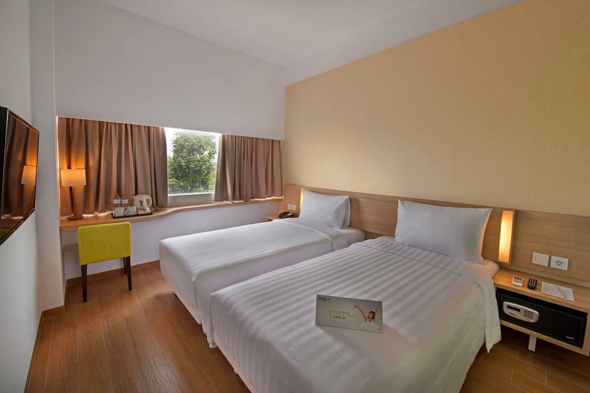 Una camera d'albergo con due letti, una scrivania con una sedia, una finestra con tende e un comodino con una lampada e un telefono.