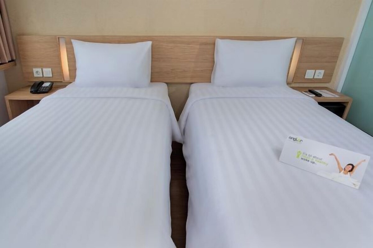 A hotel room with two single beds, each with a wooden headboard, white bedding, bedside tables with lamps, and a small card on the right bed.