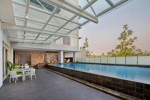 Un espace piscine extérieur moderne doté d’un toit en verre, d’une table à manger avec des chaises et d’un coin salon avec des coussins.