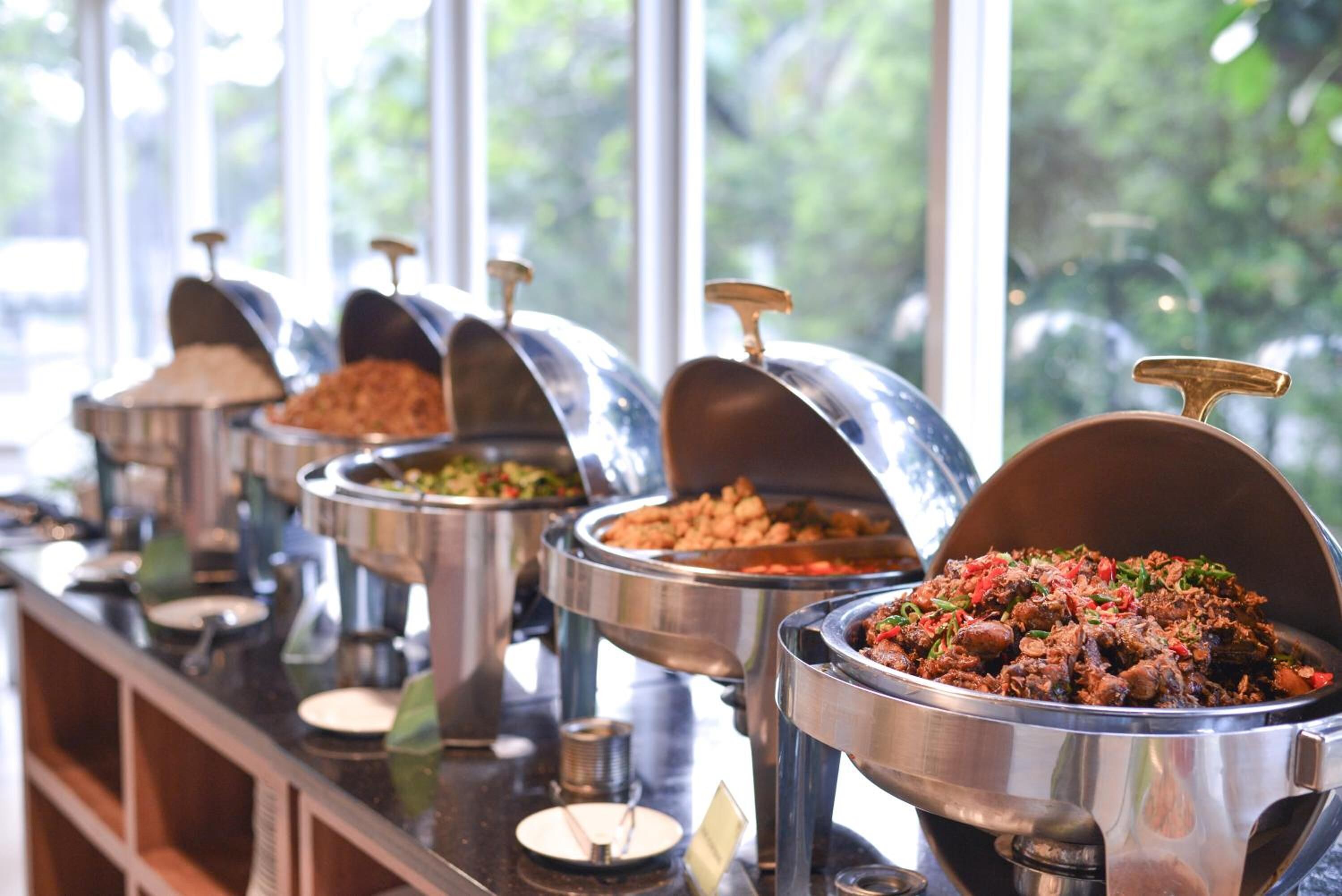 A buffet line with various food dishes in metal chafing dishes, set against a window with a view of greenery outside.