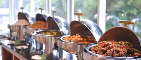 A buffet line with various food dishes in metal chafing dishes, set against a window with a view of greenery outside.