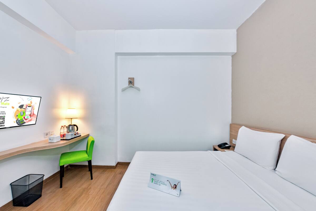 Una camera d'albergo con un letto, una scrivania con una sedia, una lampada, un cestino e una TV a parete.