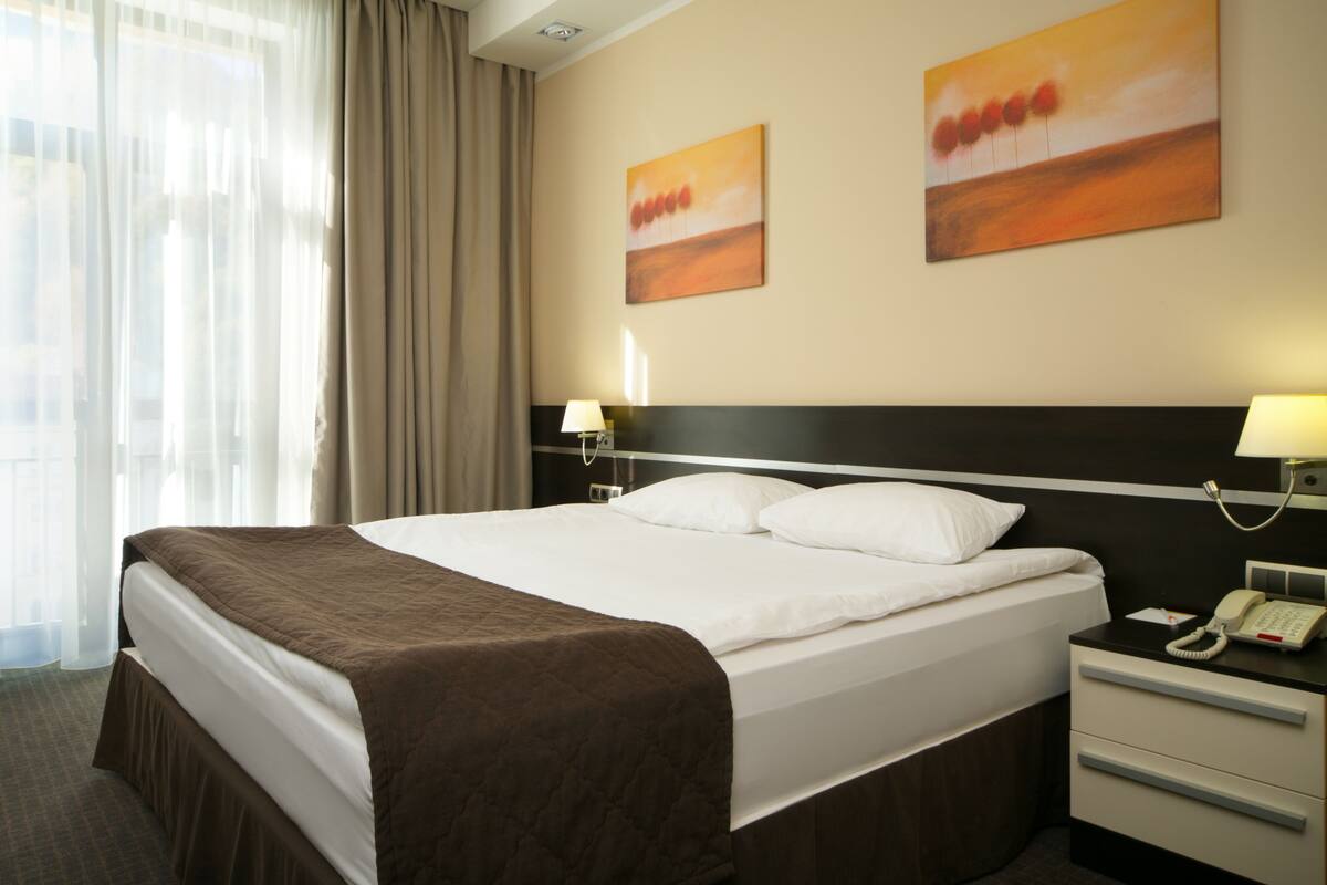 Hotellihuone, jossa on suuri sänky, yöpöytä lampun kanssa, puhelin ja kaksi taulua seinällä.