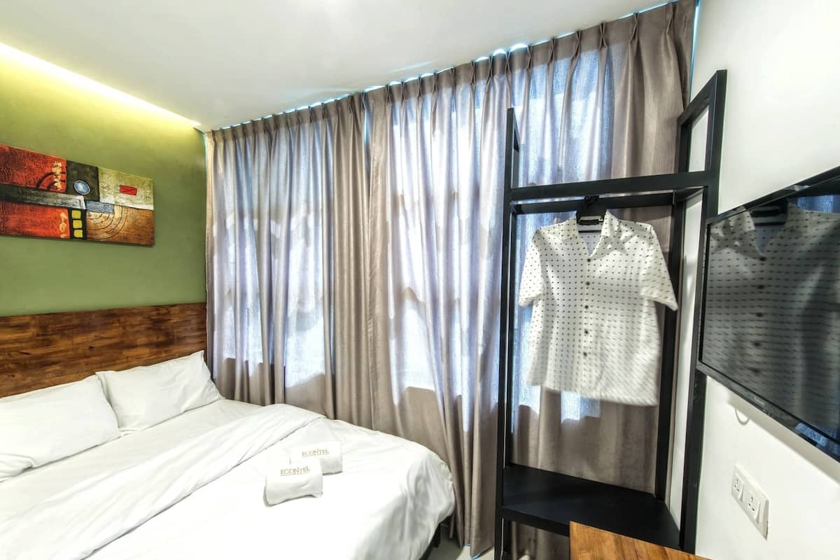 A hotel room with a bed, a wooden headboard, a window with curtains, a TV, and a wall-mounted shelf with a shirt.