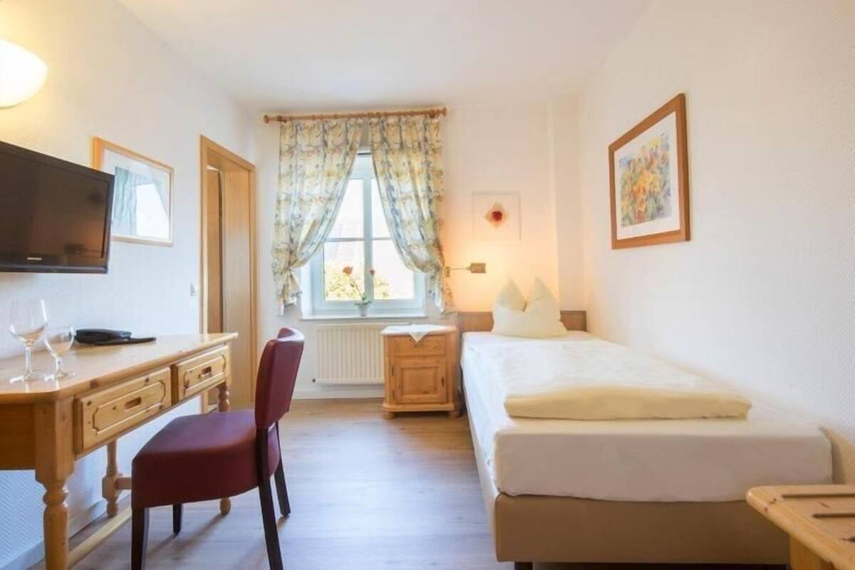 A bedroom with a bed, a desk with a chair, a television, and a window with curtains.