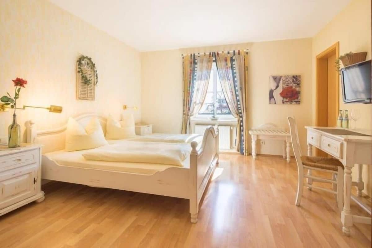 A bedroom with a bed, a desk, a chair, a TV, and a window with curtains.