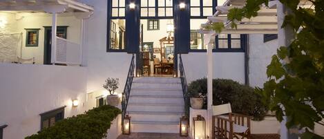 A white building with blue accents, a staircase leading to a second floor, stone flooring, and outdoor seating area with lanterns.