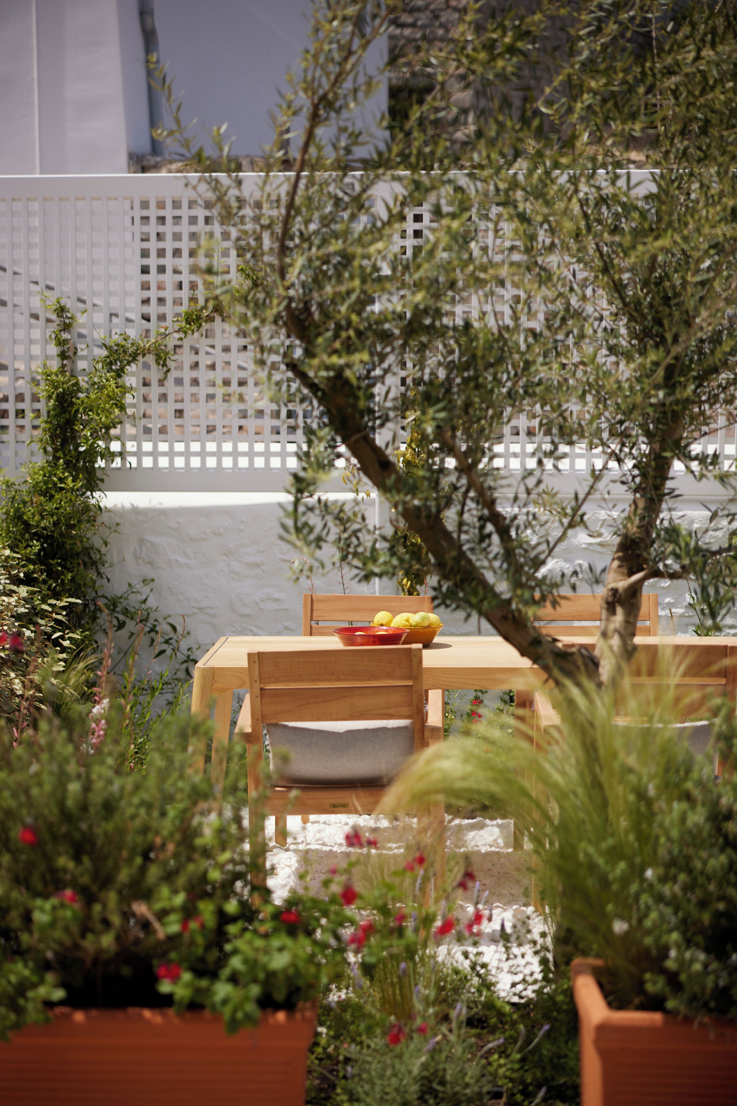 A wooden table with a bowl of fruit and a chair in a garden area with potted plants and a white fence in the background.
