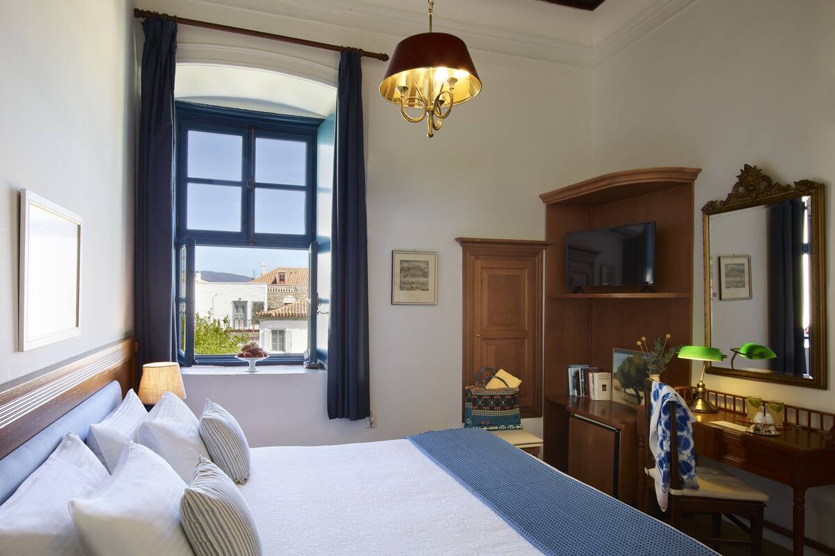 Habitación de hotel con una cama grande, un escritorio con televisor, una ventana con cortinas y vistas a edificios.