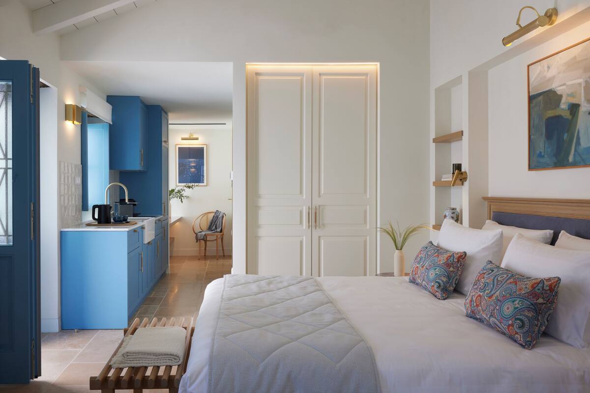 Un dormitorio con una cama grande, una mesita de noche y una lámpara auxiliar. Hay una ventana con cortinas, una lámpara de noche y un estante en la pared.
