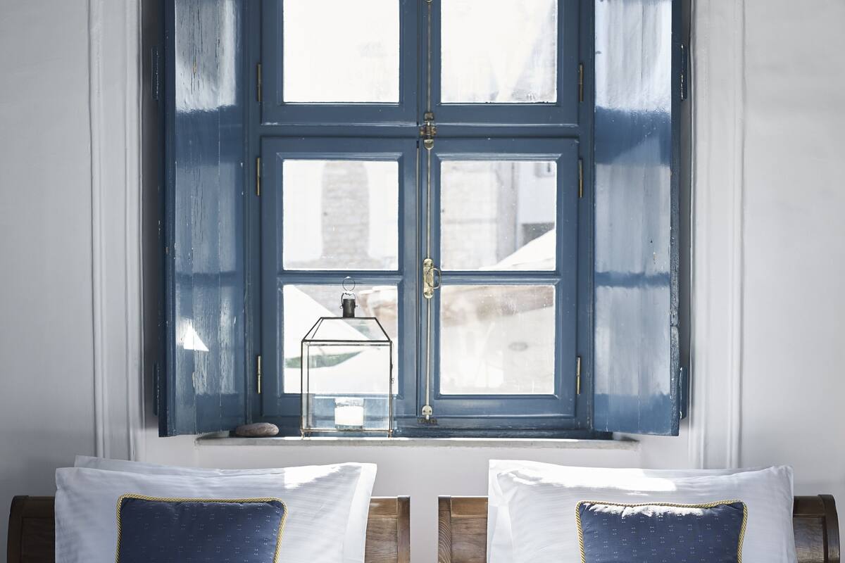 Una cama con ropa de cama blanca y dos almohadas azules, una ventana con contraventanas azules y un pequeño farol en el alféizar.