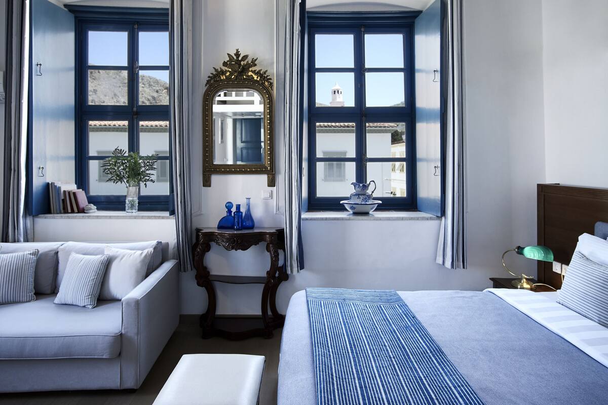 Habitación de hotel con cama, sofá, mesita de consola con objetos decorativos y vistas a una montaña y un edificio blanco a través de dos ventanas.