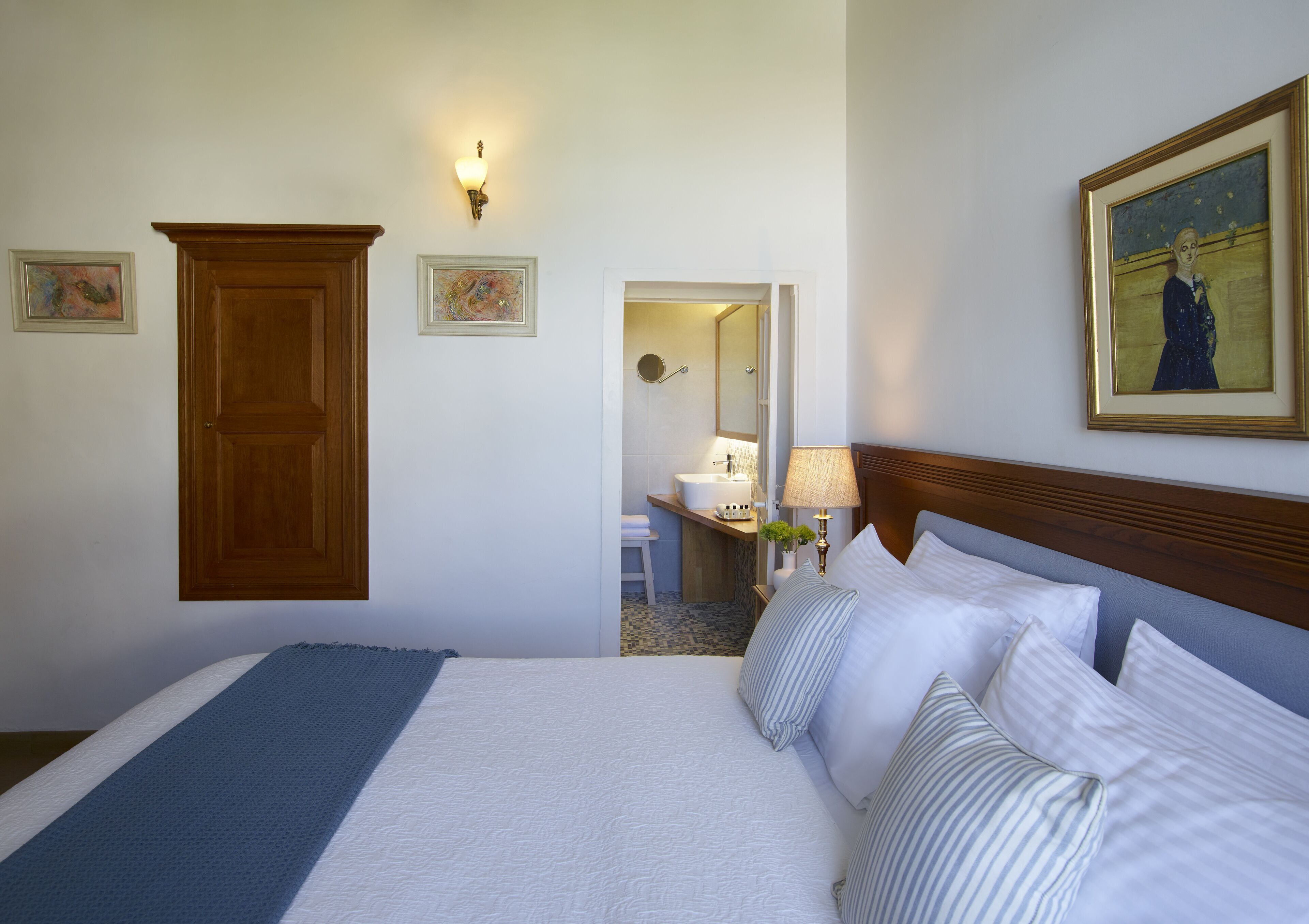 A hotel room with a large bed, a wooden wardrobe, a bathroom visible through an open door, and a painting on the wall.