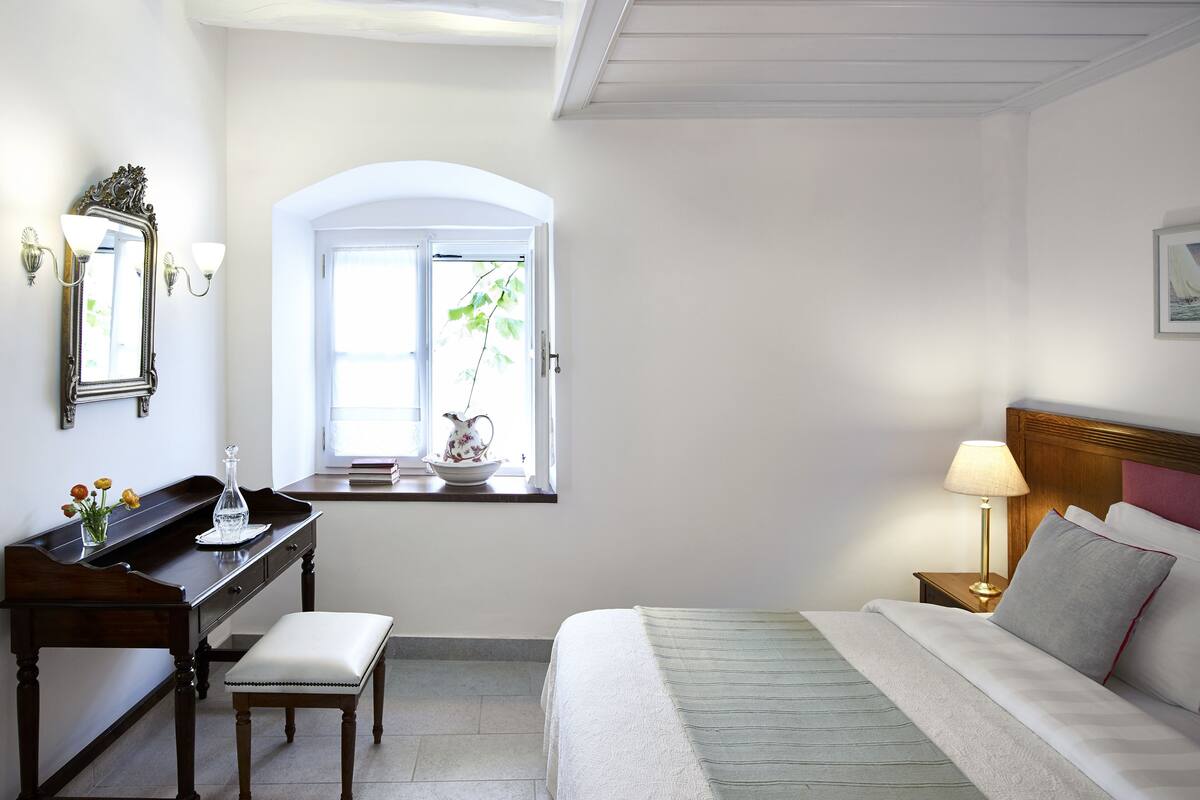 Un dormitorio con cama, mesita de noche, lámpara y ventana con vistas a la vegetación.