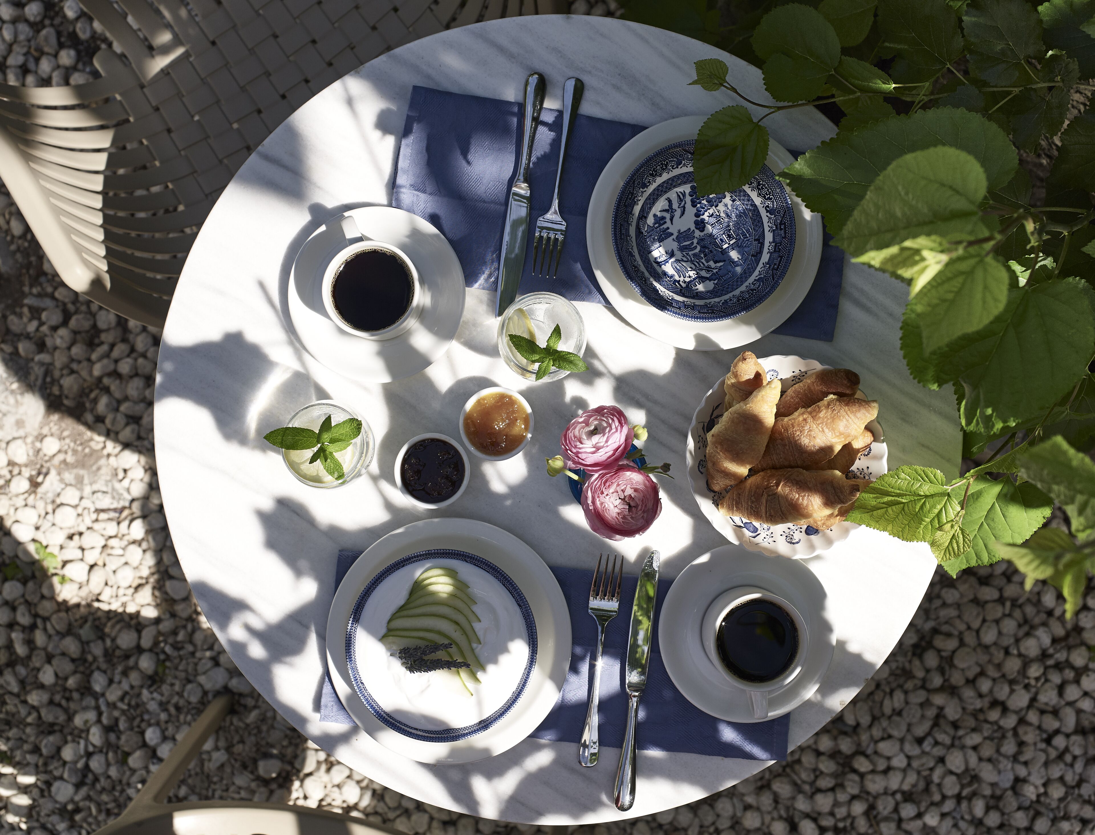 Una mesa redonda con mantel blanco y azul, dispuesta con platos, tazas, un tenedor, un cuchillo y alimentos como manzanas, pan y un pequeño recipiente de mermelada.