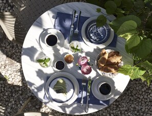 Ein runder Tisch mit weiß-blauer Tischdecke, gedeckt mit Tellern, Tassen, Gabel, Messer und Speisen wie Äpfeln, Brot und einer kleinen Schale Marmelade.