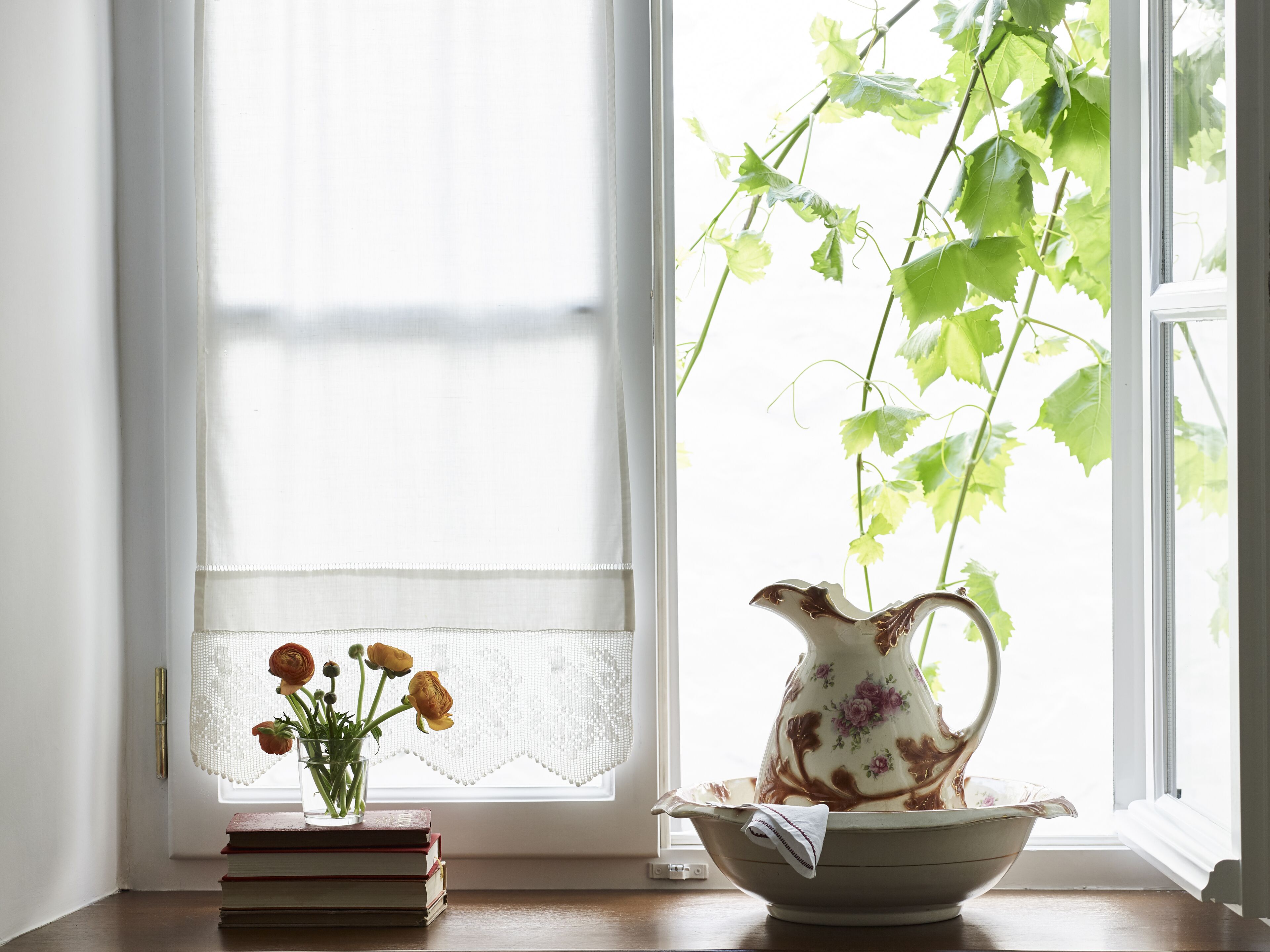 A window with a lace curtain, a vase with flowers, and a pitcher on a table.