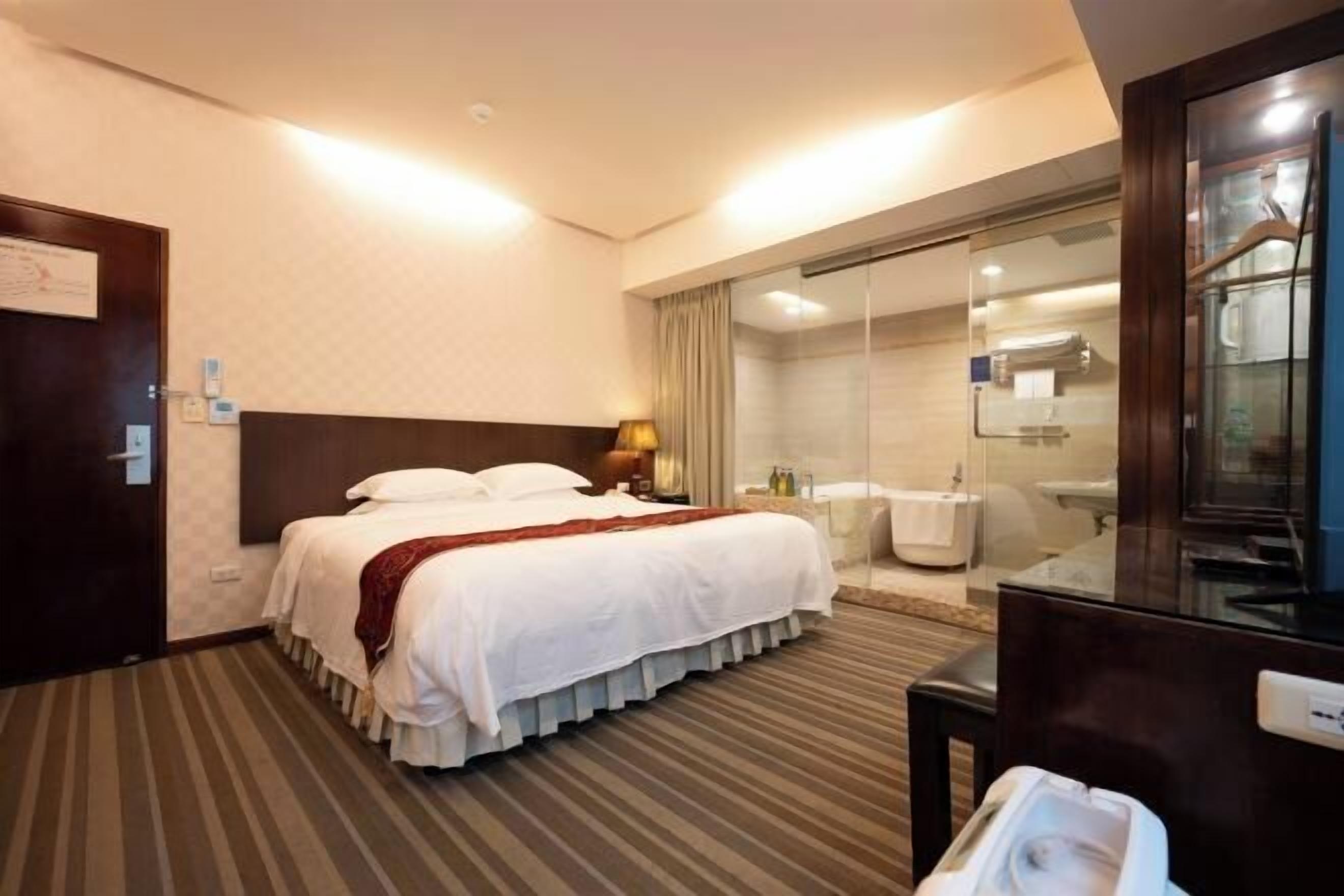 Une chambre d’hôtel comprenant un lit, un bureau, un miroir, une salle de bain avec douche et une porte en verre donnant accès à la salle de bain.