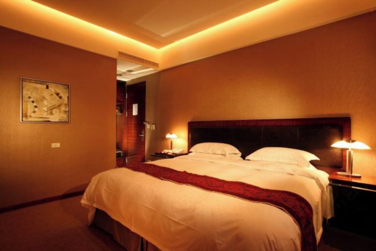 A hotel room with a large bed, two bedside lamps, a red throw blanket, and a painting on the wall.