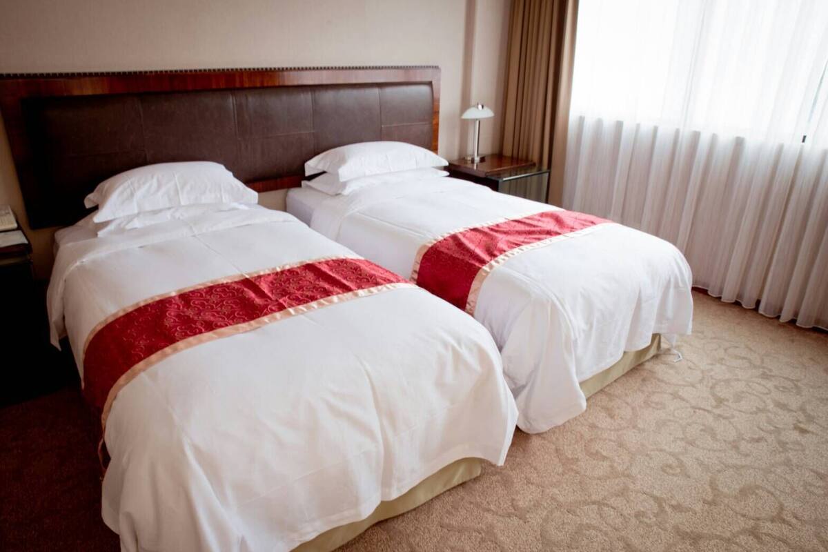 A hotel room with two beds, a wooden headboard, a nightstand with a lamp, and a window with curtains.