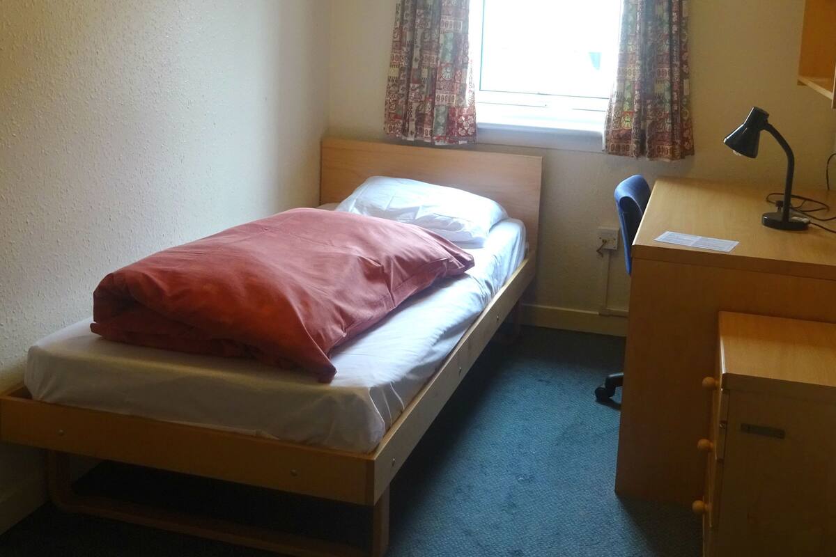 A small, single-bed room with a wooden bed frame, a desk with a lamp, and a window with patterned curtains.