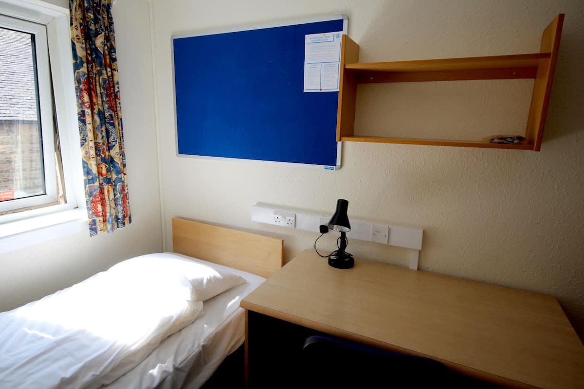 A small, tidy room with a single bed, a desk with a lamp, a bulletin board, and a window with patterned curtains.