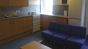 A compact kitchen with wooden cabinets, a stove, a sink, and a refrigerator. There is a window providing natural light and a blue chair in the room. - Edinburgh Metro Youth Hostel (Edinburgh)