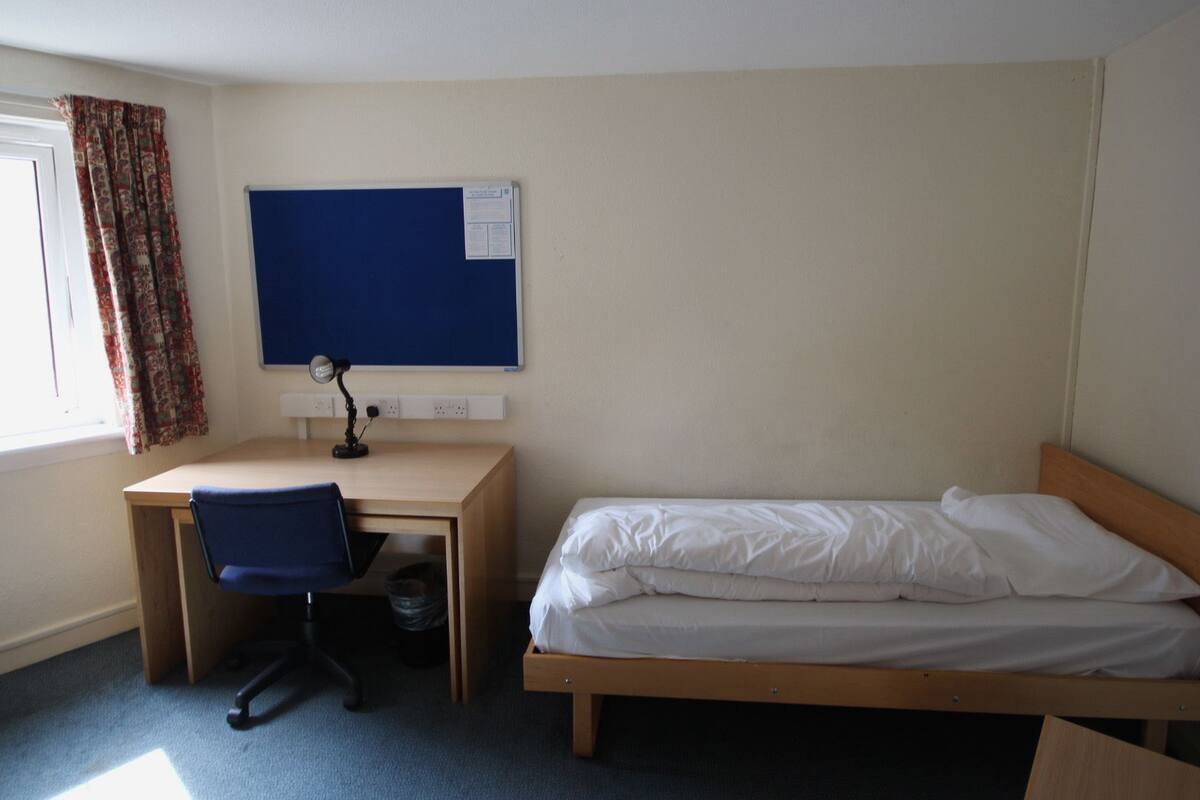 A room with a single bed, a desk with a chair, a bulletin board, and a window with curtains.