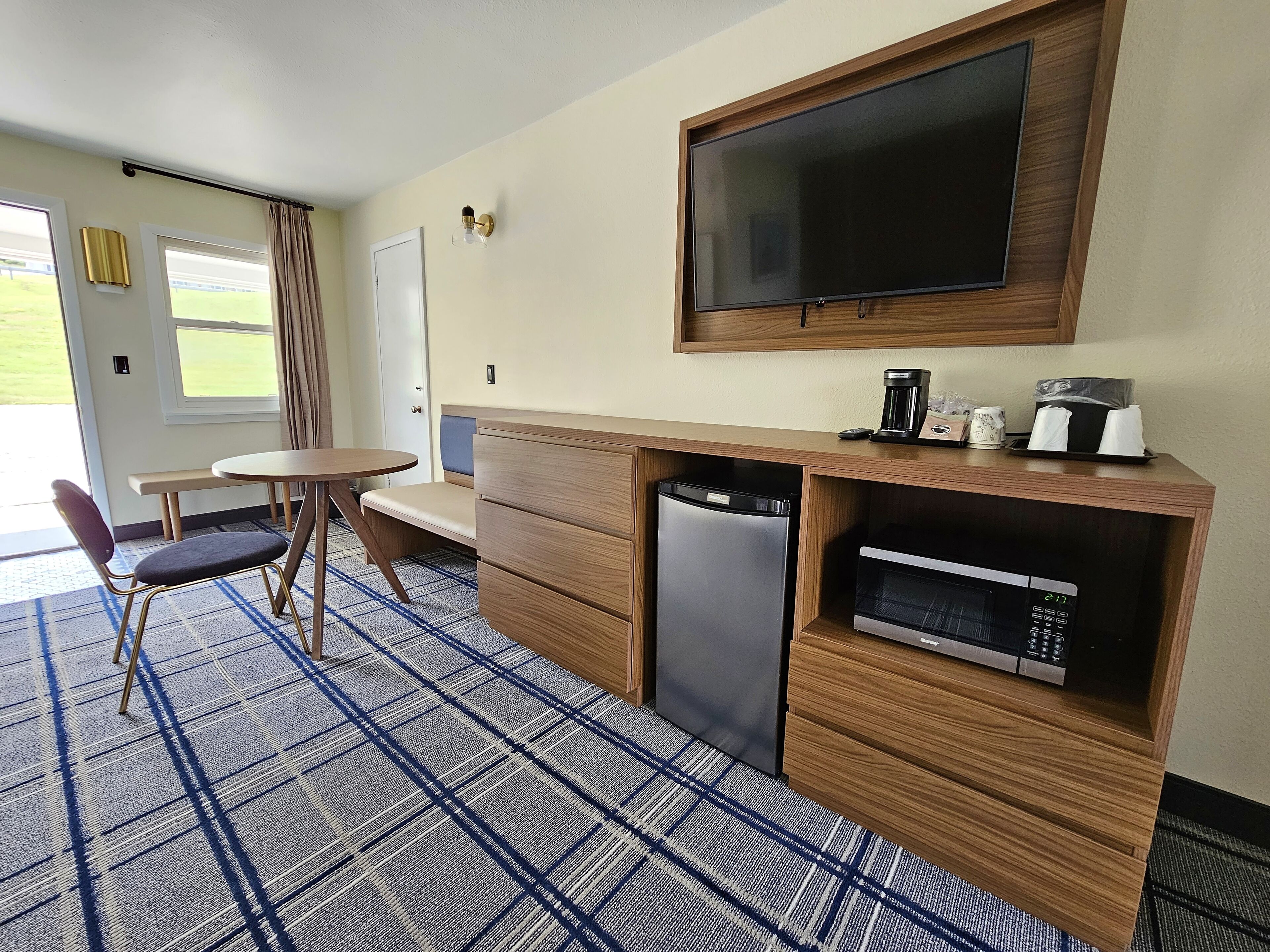 A compact hotel room with a kitchenette, a flat-screen TV mounted above the counter, a small dining table with a chair, and a large window offering a view of the outdoors.