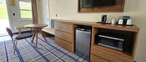 Ett kompakt hotellrum med ett litet pentry, en platt-TV som är monterad ovanför disken, ett litet matbord med en stol och ett stort fönster med utsikt över omgivningarna.