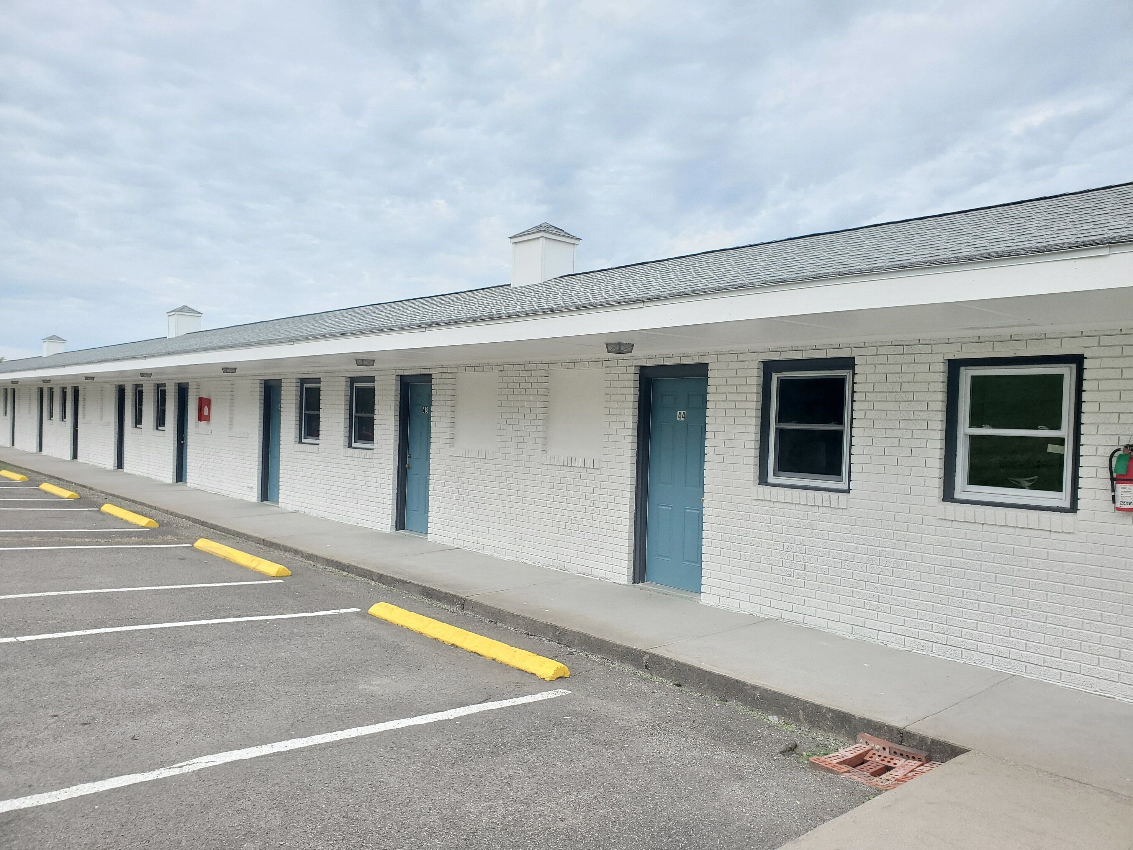 Una hilera de habitaciones de motel con puertas individuales, un estacionamiento con espacios delimitados y cielo despejado.