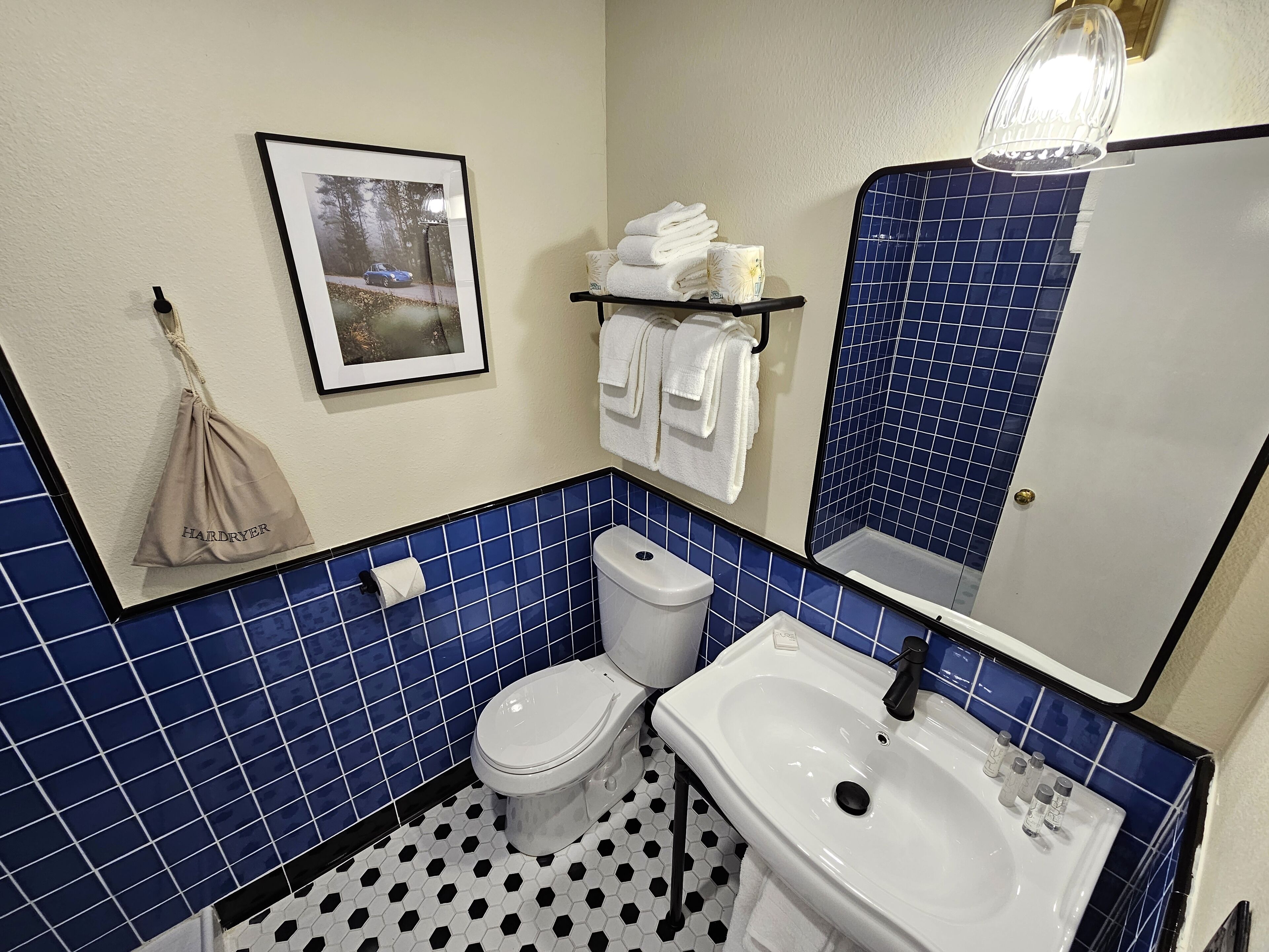 A bathroom with a toilet, sink, and blue-tiled walls. There is a framed picture on the wall, a towel rack with towels, and a hanging bag labeled "Hardryer".